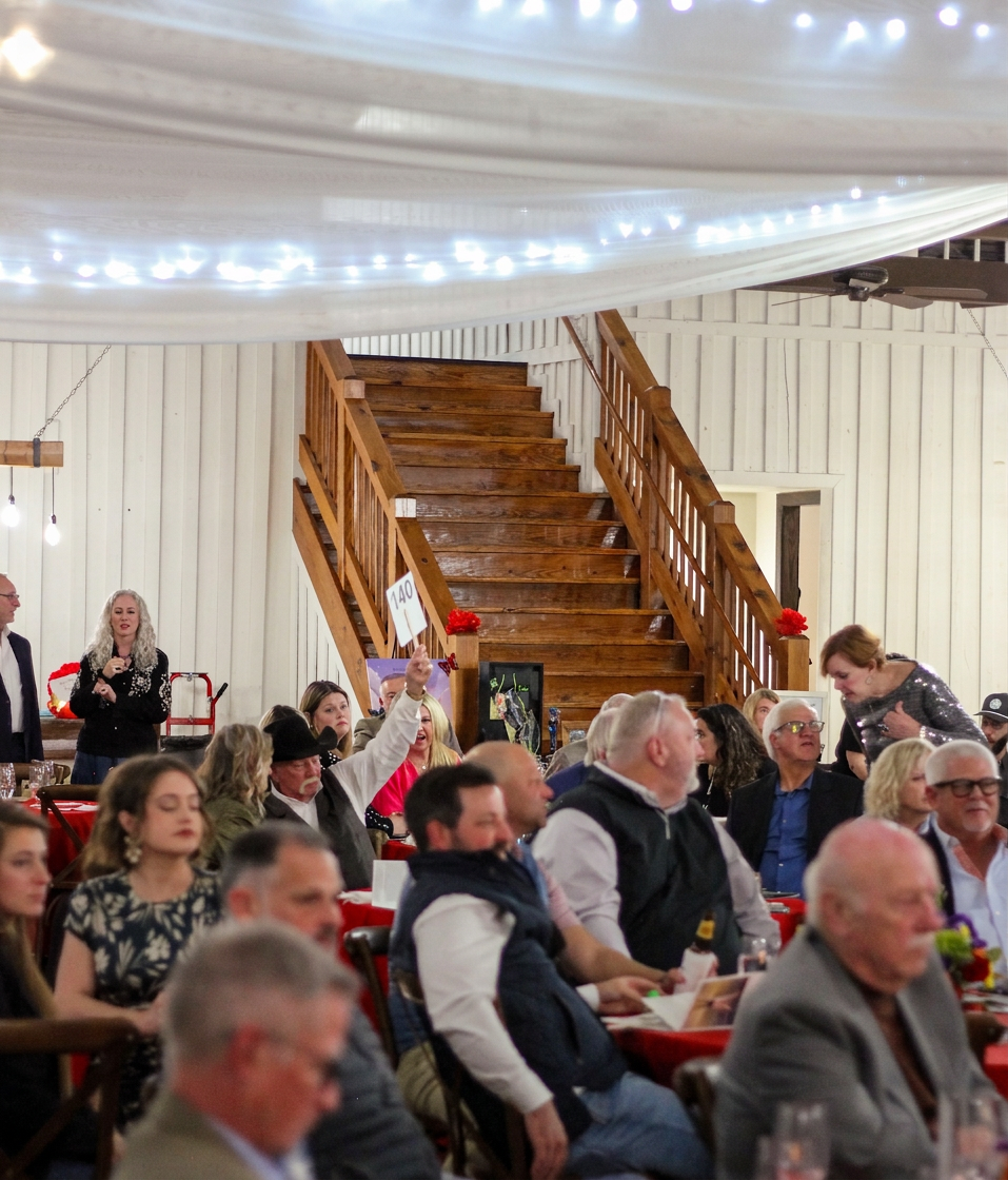Guests participate in a live charity auction at the Friends of Vail Foundation’s annual Dinner With Friends fundraising event in North Texas. Attendees gather beneath string lights at a rustic venue, raising bid paddles to support childhood literacy, education, scholarships, and community programs.
